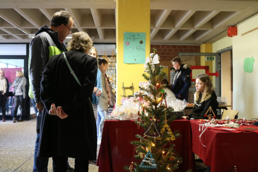 Weihnachtsmarkt mit Ständen und Besuchern in einer Schule. Ein Baum ist geschmückt.