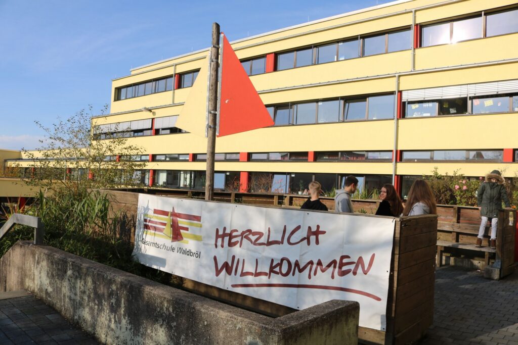 Schuleingang mit Willkommensbanner und Flagge, moderne Gebäude im Hintergrund.