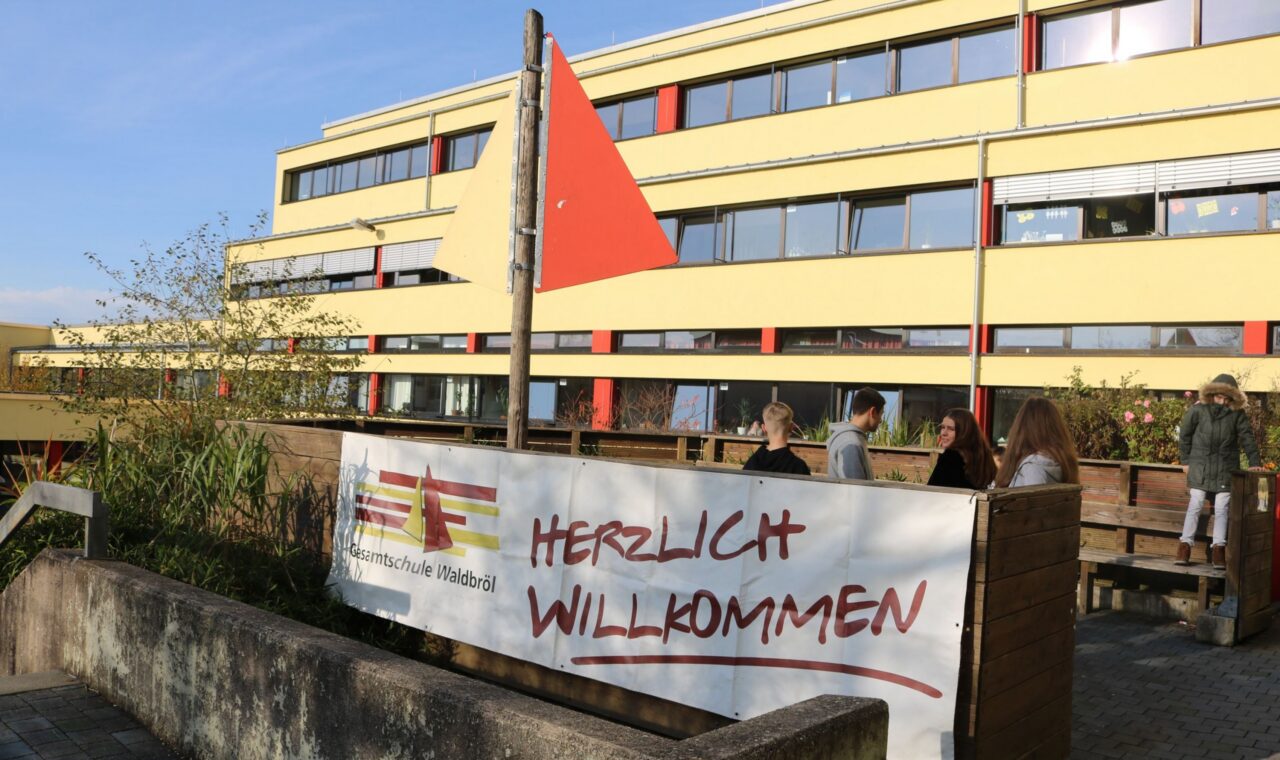 Schuleingang mit Willkommensbanner und Flagge, moderne Gebäude im Hintergrund.