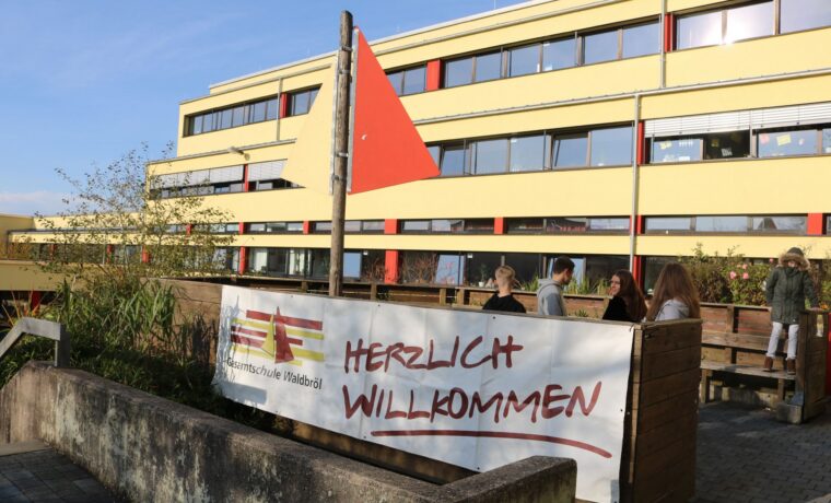 Schuleingang mit Willkommensbanner und Flagge, moderne Gebäude im Hintergrund.