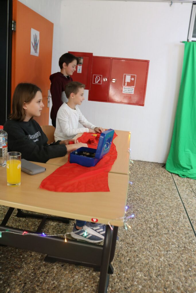 Kinder sitzen an einem Tisch vor einem grünen Hintergrund und bereiten etwas vor.