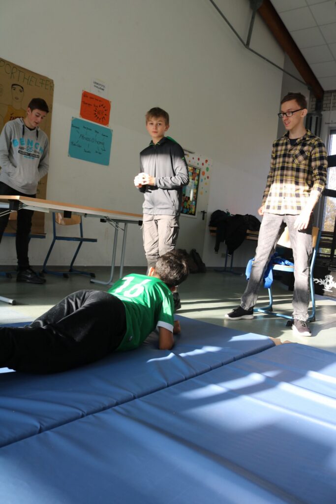 Schüler liegen auf Matten, während andere sie beobachten und an einem Tisch stehen.