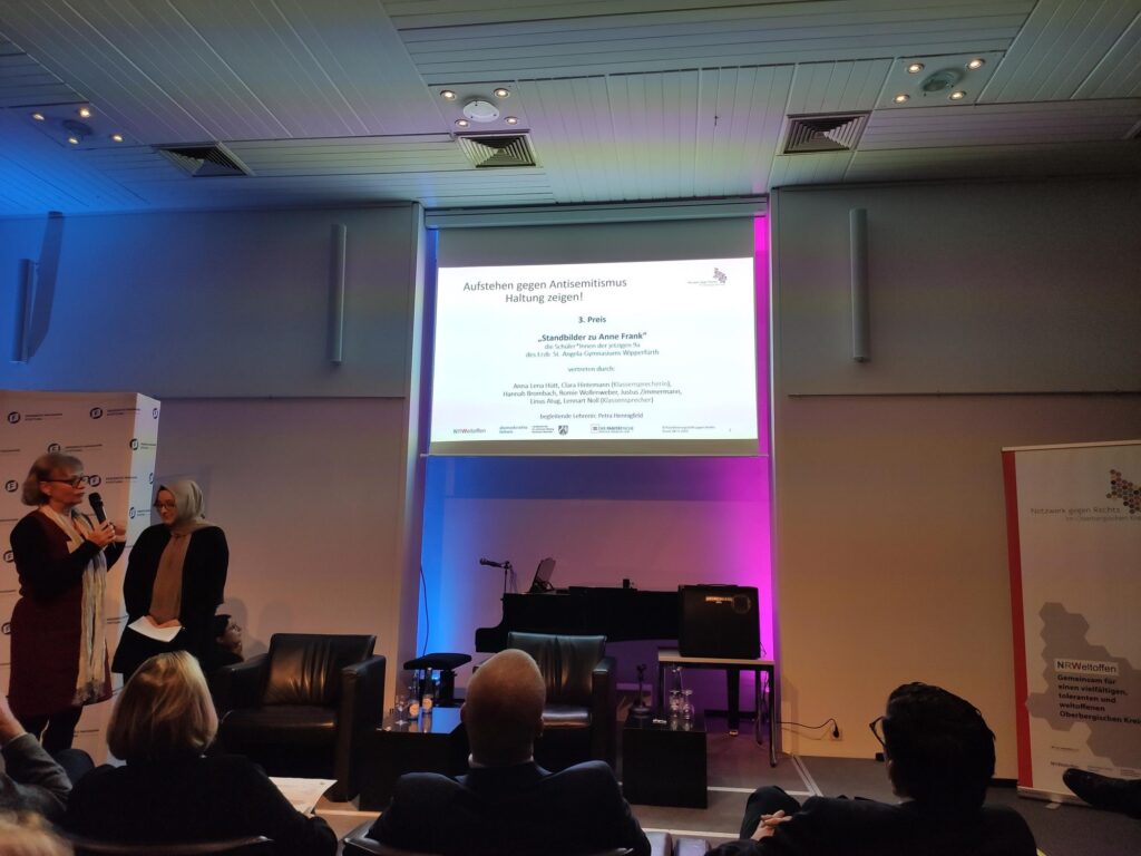 Veranstaltungssaal mit Podium und Publikum, Präsentation auf großer Leinwand.