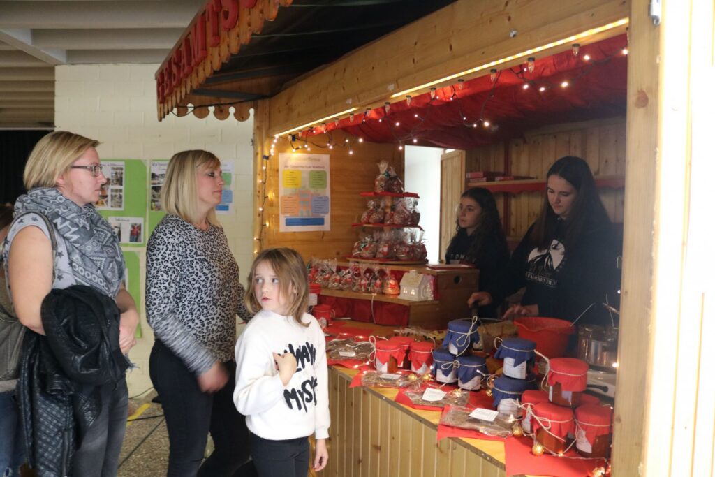Marktstand mit Süßigkeiten, Besucherinnen und einem Kind. Verkaufsaktivität im Hintergrund.