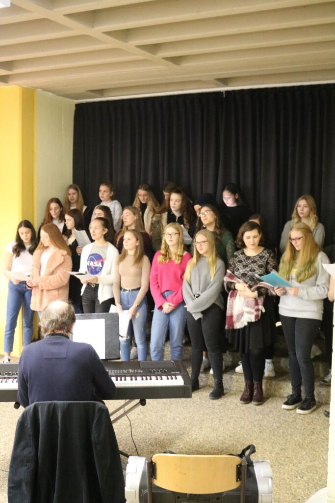 Chor mit Sängerinnen vor einem Klavier in einem Probenraum.