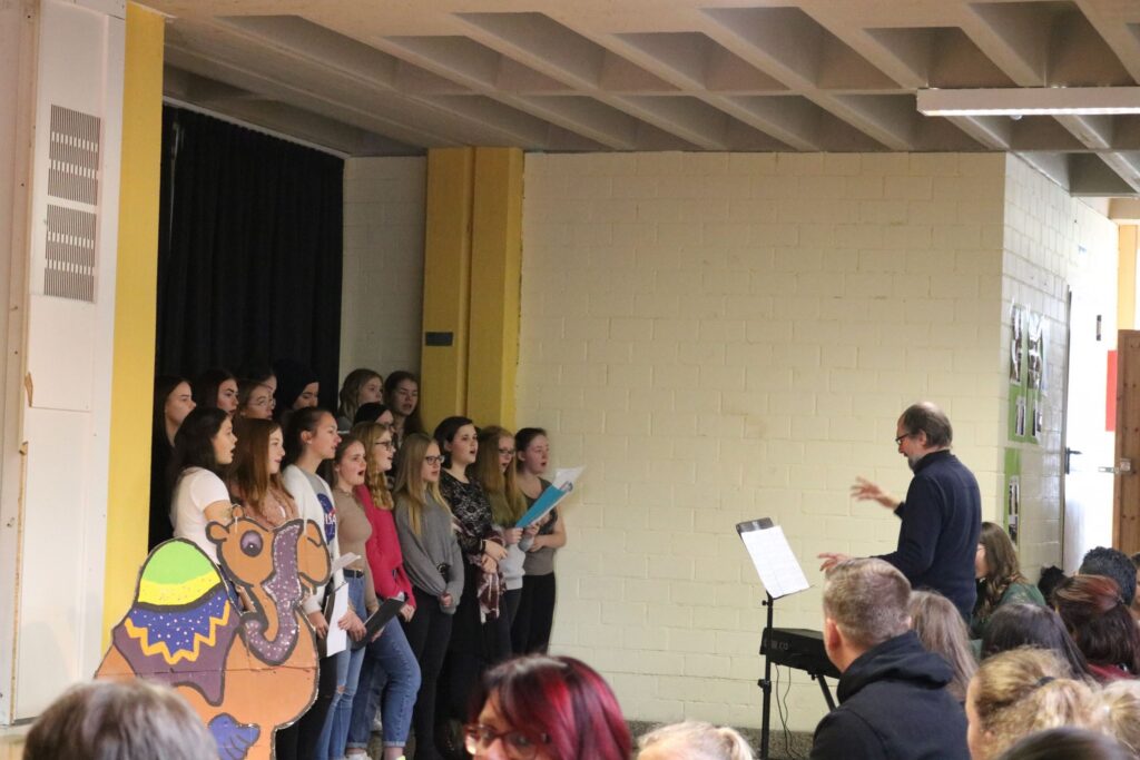 Choraufführung in der Schule mit Lehrer und Schülerinnen auf der Bühne.