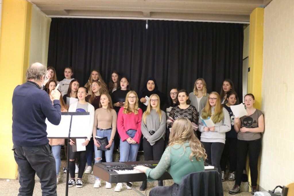 Chorprobe mit Schülerinnen und Lehrer am Klavier in einem Raum.