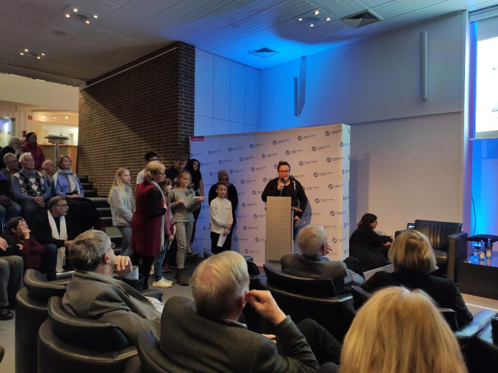 Veranstaltung mit Vortrag und Publikum im Hintergrund, Bühne im Vordergrund.