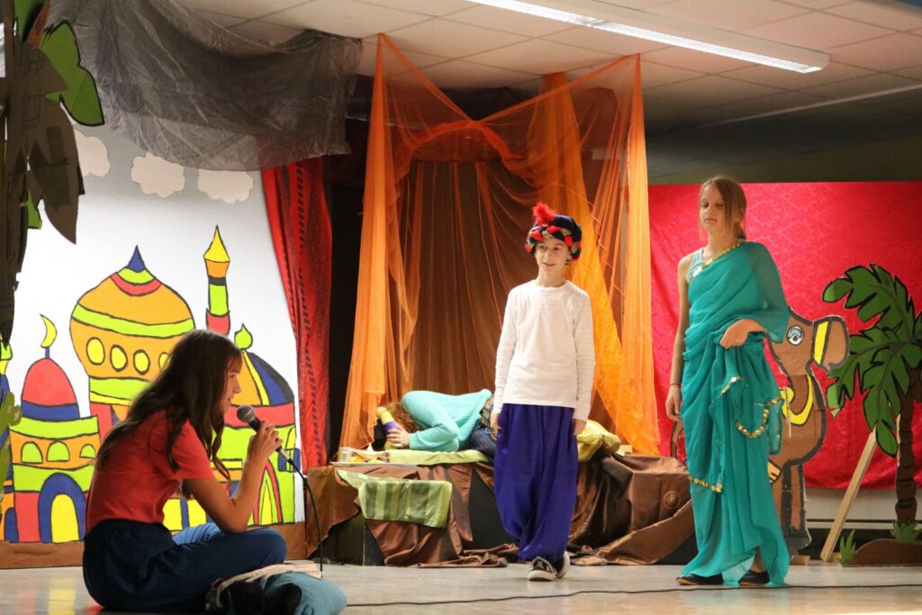 Zwei Kinder in Kostümen auf einer Theaterbühne; eine Kulisse mit bunten Gebäuden.