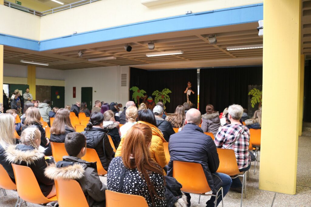 Schüler stehen auf einer Bühne, Publikum sitzt in orangefarbenen Stühlen.