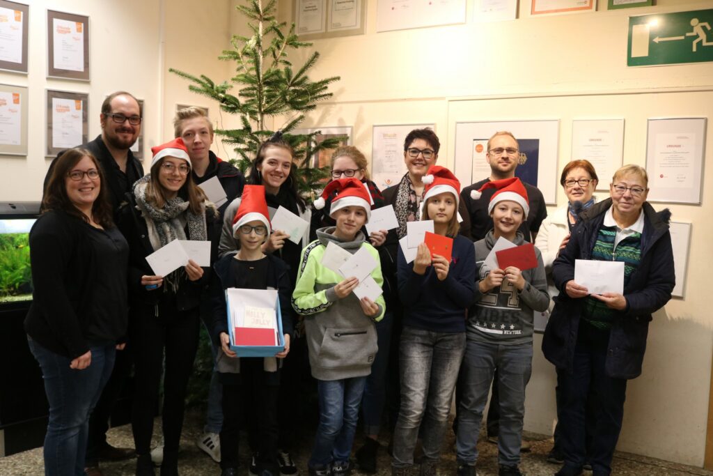 Gruppe von Personen mit Weihnachtsmützen, die Karten halten, vor Weihnachtsbaum.