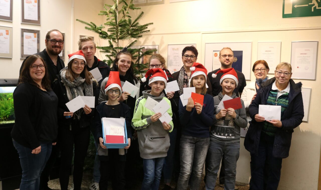 Gruppe von Personen mit Weihnachtsmützen, die Karten halten, vor Weihnachtsbaum.