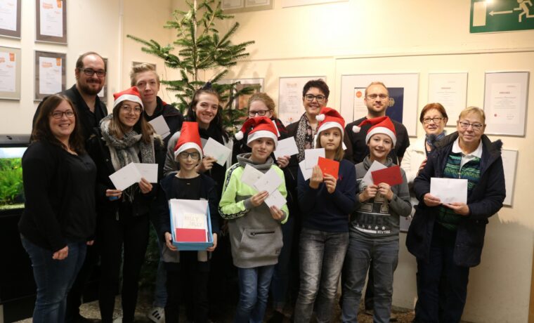 Gruppe von Personen mit Weihnachtsmützen, die Karten halten, vor Weihnachtsbaum.