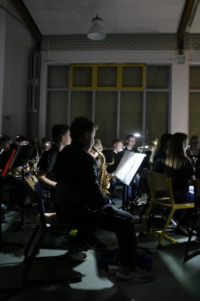 Schüler bei einem Konzert, sitzend mit Instrumenten und Notenständern in dunklem Raum.