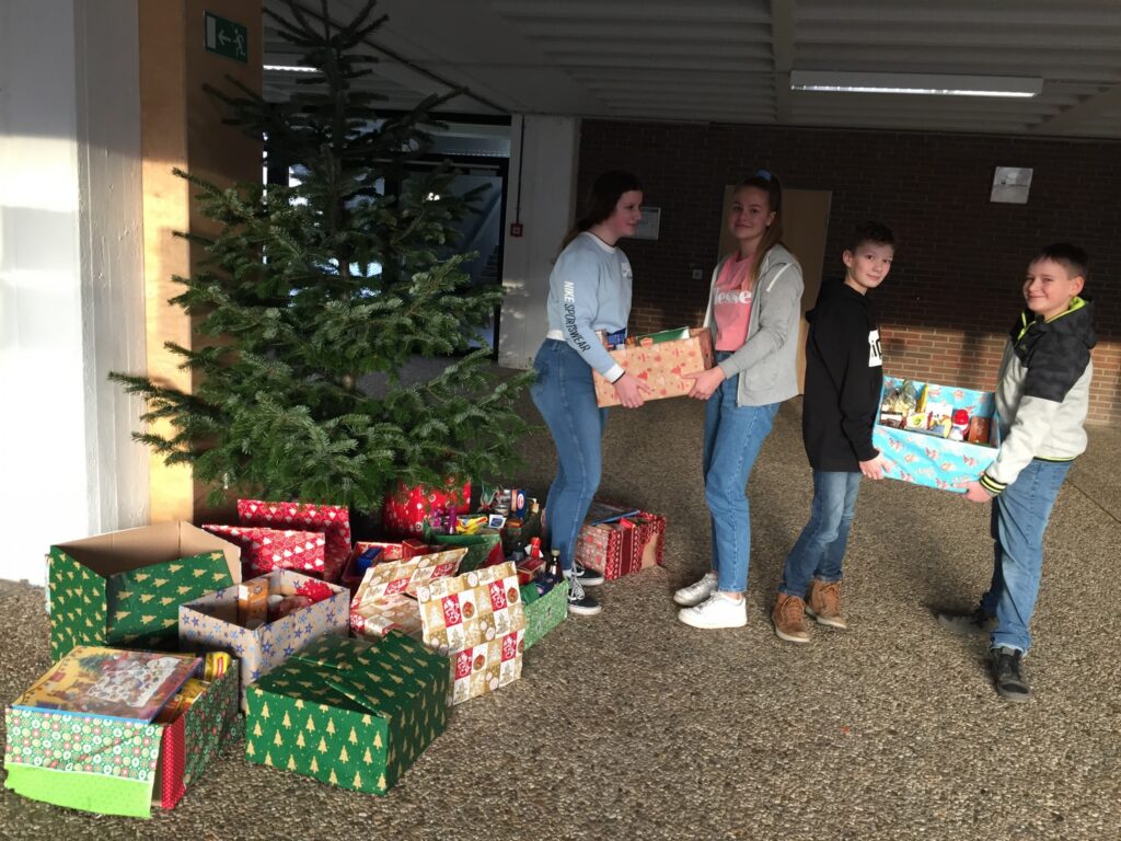 Schüler tragen Geschenke vor einem Weihnachtsbaum in einem Schulgebäude.