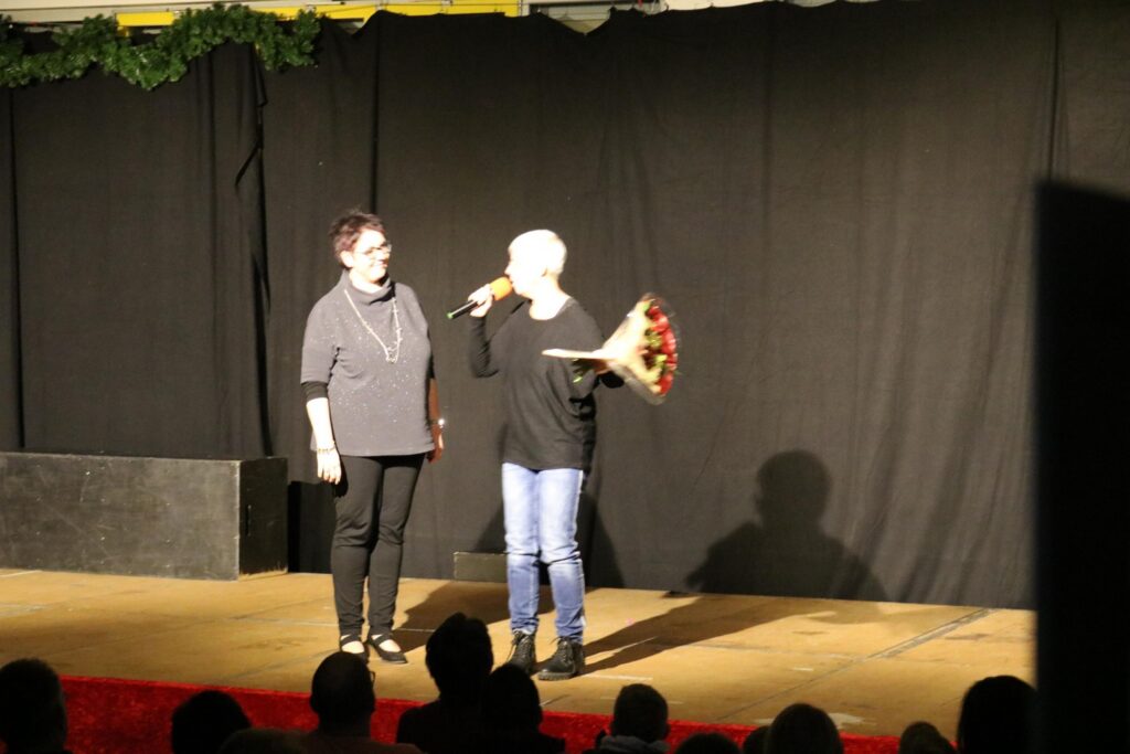 Zwei Personen stehen auf einer Bühne und sprechen. Eine hält einen Blumenstrauß.