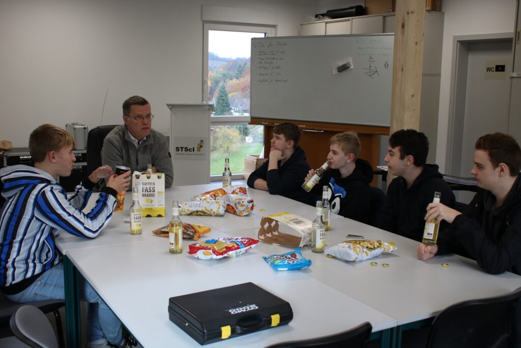 Gruppe von Jugendlichen und einem Erwachsenen sitzt an einem Tisch mit Snacks und Getränken.