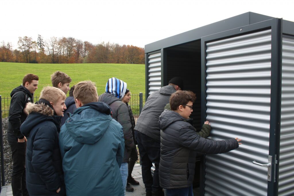 Gruppe von Schülern steht vor einem grauen, geschlossenen Container.