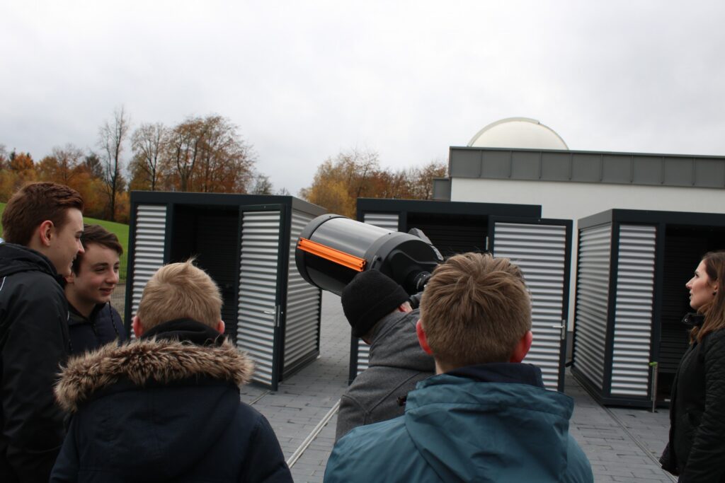 Gruppe von jungen Menschen betrachtet ein Teleskop vor einem Observatorium.