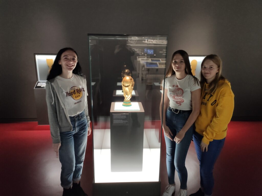 Drei Schülerinnen stehen vor einer Vitrine mit einer goldenen Statue.