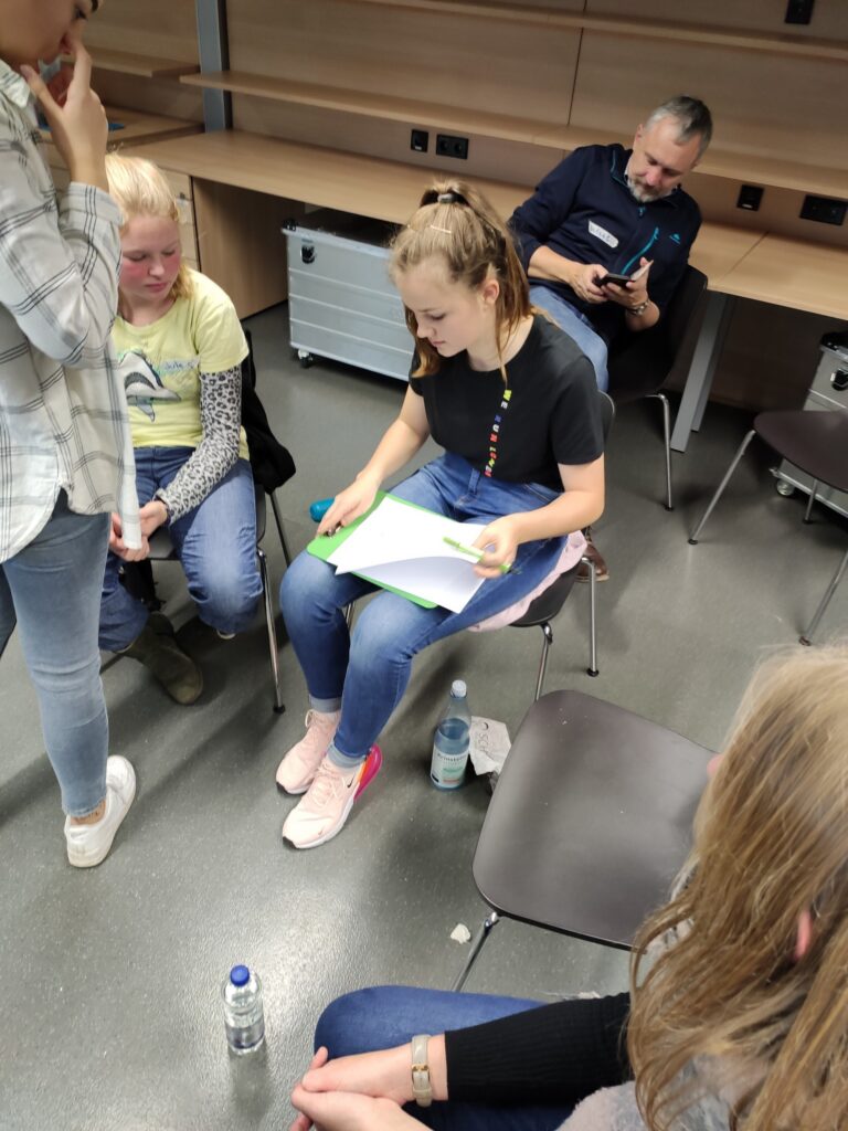 Eine Gruppe von Schülern sitzt in einem Raum. Eine Schülerin liest ein Blatt Papier.