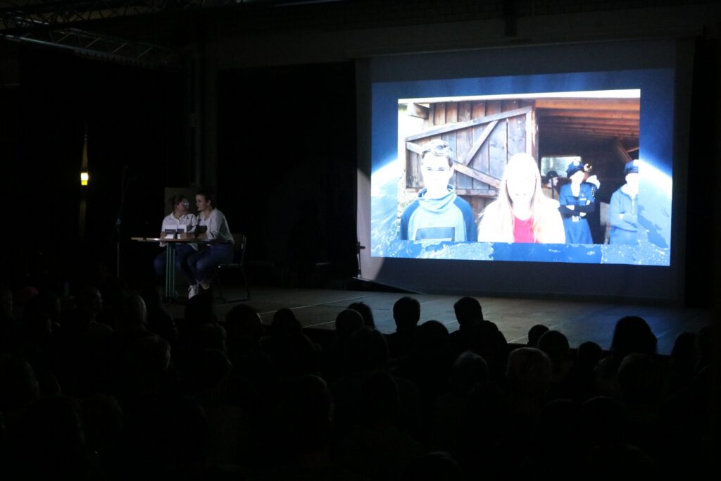 Blick auf eine Bühne mit Präsentation und Publikum im Hintergrund.