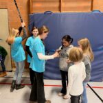 Gruppe von Kindern in blauen T-Shirts, die um eine Matte herum stehen und aktiv miteinander sprechen.