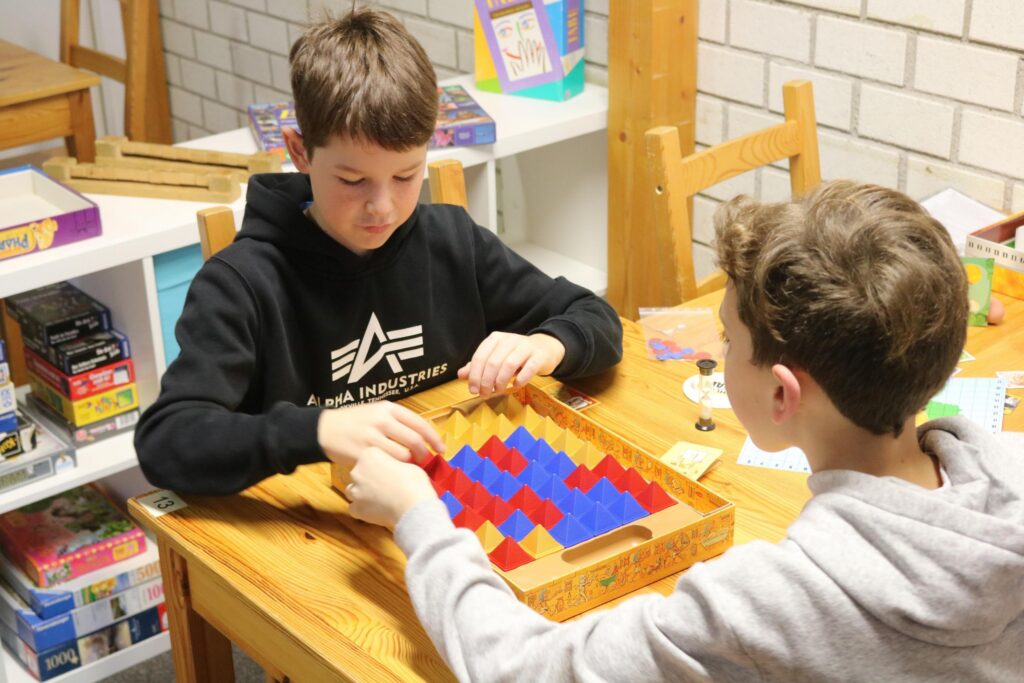 Zwei Schüler spielen ein Brettspiel am Tisch in einem Klassenraum.