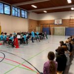 Schulkonzert mit Schülern in blauen T-Shirts, Musikern auf der Bühne und Publikum.