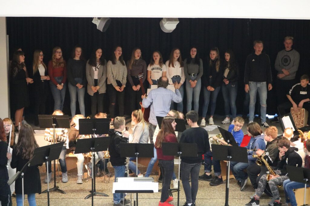 Schüler*innen stehen auf der Bühne, während andere im Orchester spielen.