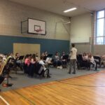 Musikgruppe probt in einer Sporthalle mit Instructor. Notenständer und Instrumente sichtbar.