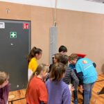 Eine Gruppe von Kindern steht in einer Turnhalle und blickt auf einen anderen, der etwas erklärt.