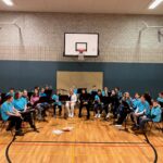 Probe einer Schulband in einer Turnhalle mit Instrumenten und Spielern in blauen T-Shirts.