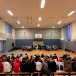 Schüler sitzen in einem Schulraum, während andere ein Konzert auf der Bühne geben.