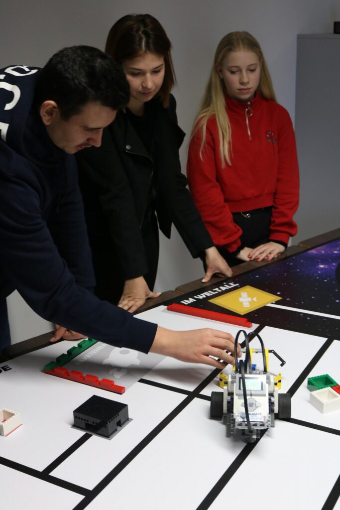 Eine Person zeigt zwei Schülerinnen einen Roboter auf einem Tisch.