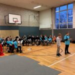 Schüler stehen auf einer Sporthalle vor einer Band und präsentieren ein Programm.