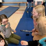 Kinder sitzen auf Turnmatten und unterhalten sich, eine hat ein Instrument in der Mitte.