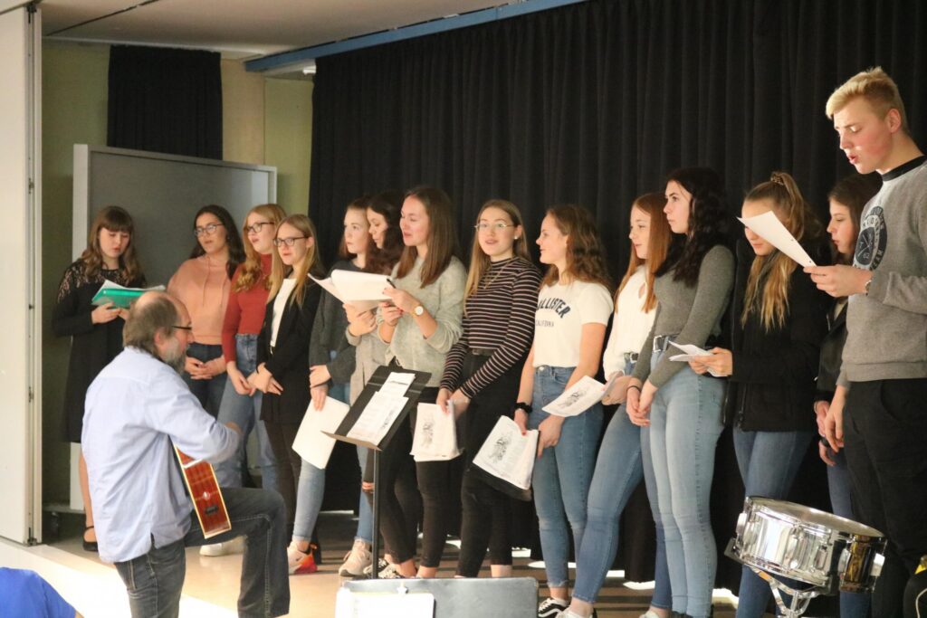 Schüler singen im Schulgebäude, begleitet von einem Gitarristen.