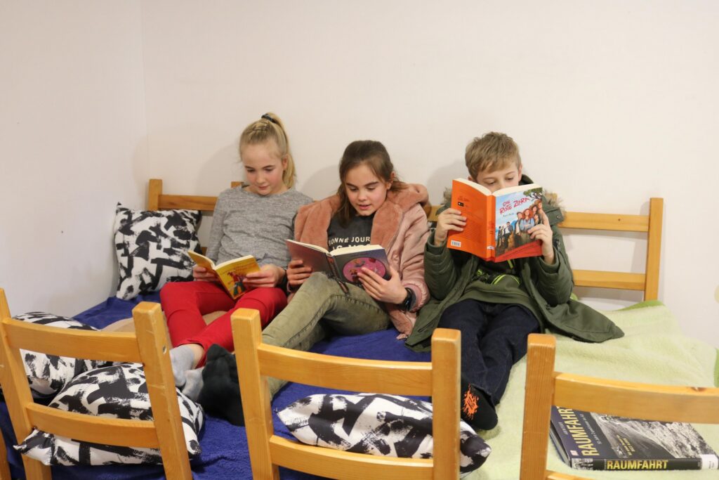 Drei Kinder sitzen auf einem Bett und lesen Bücher.