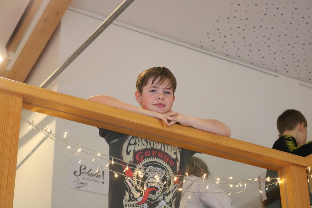 Junge lehnt an einem Geländer und schaut in die Kamera. Im Hintergrund sind andere Kinder.