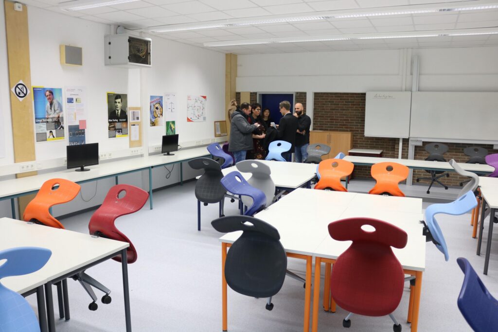 Leerer Klassenraum mit bunten Stühlen und Tischen. Schüler stehen zusammen.