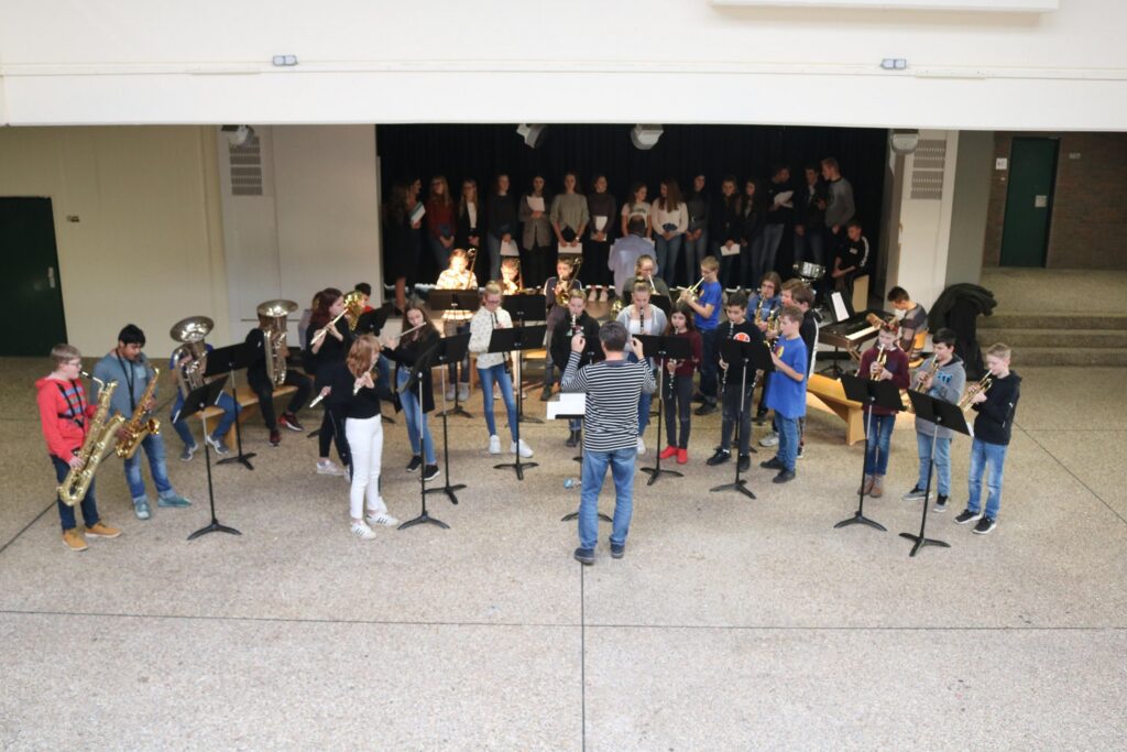 Schüler spielen im Schulgebäude gemeinsam Musik, dirigiert von einer Lehrkraft.