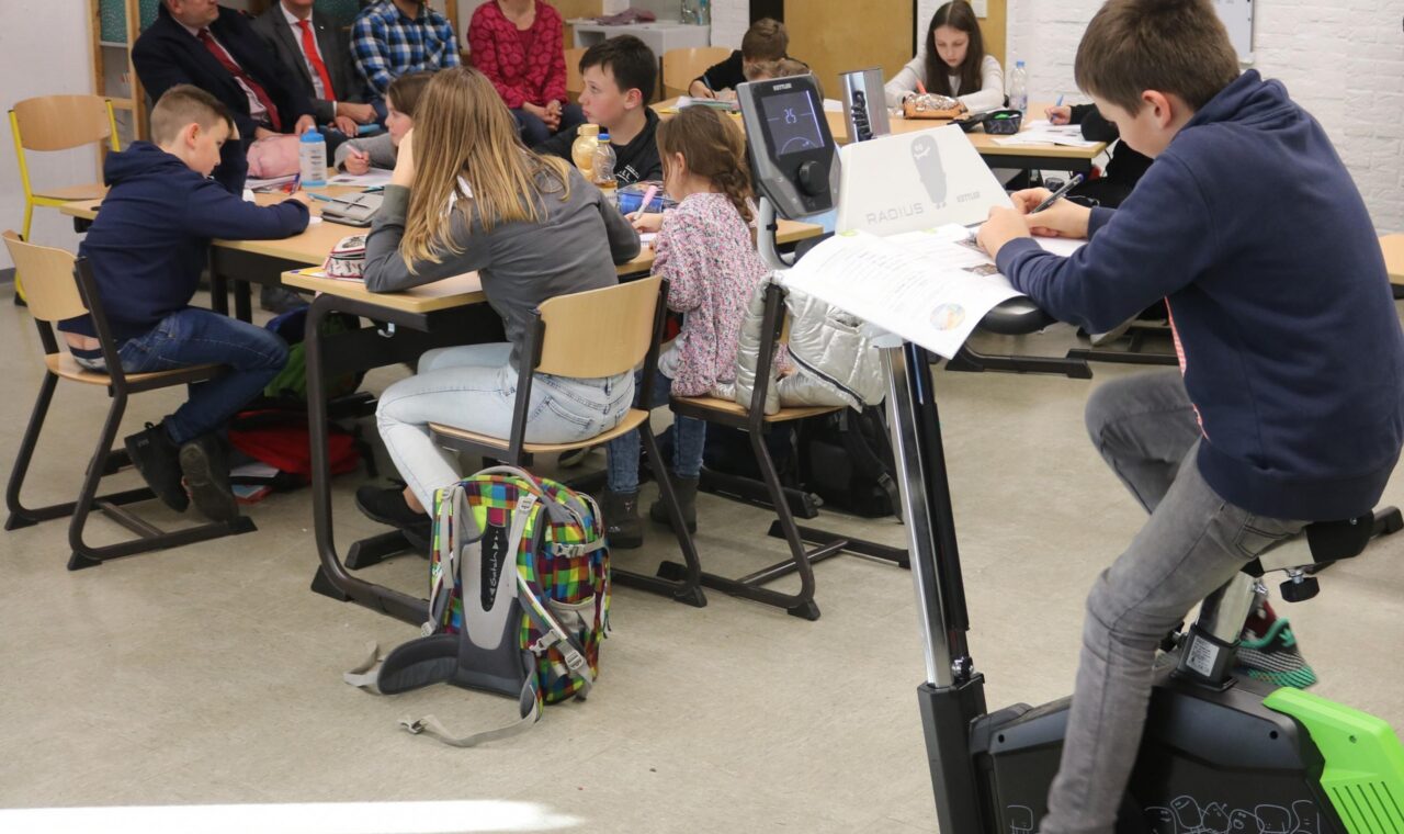 Schüler sitzen an Tischen, ein Junge fährt auf einem Fitnessgerät.