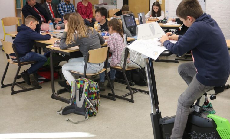 Schüler sitzen an Tischen, ein Junge fährt auf einem Fitnessgerät.