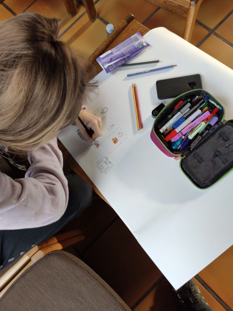 Ein Mädchen zeichnet auf einem großen Blatt Papier, umgeben von Stiften und einem Handy.