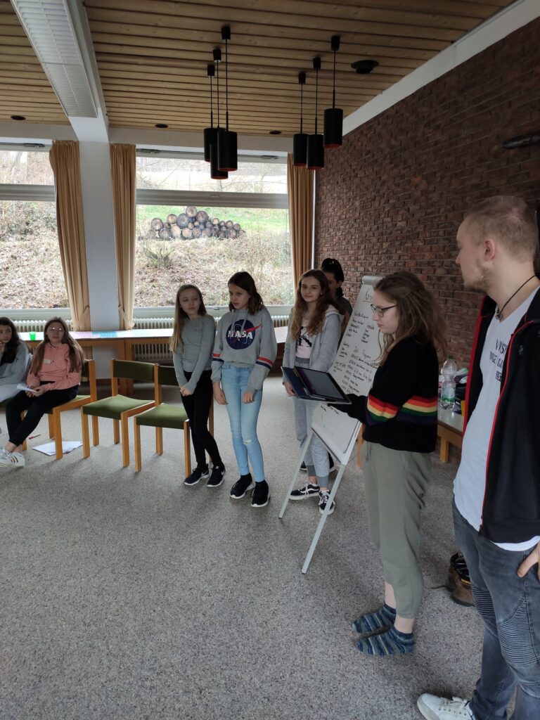Gruppe von Jugendlichen steht in einem Raum, Präsentation vor einem Flipchart.