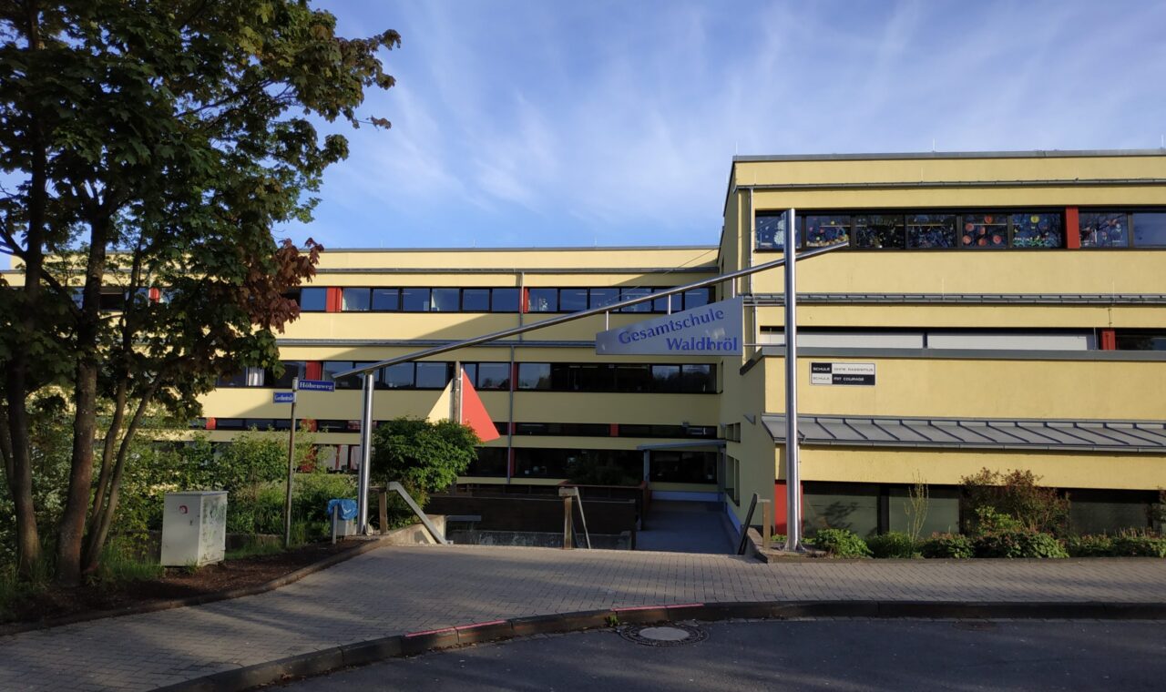 Schulgebäude mit Eingang und Flagge im Vordergrund, blauer Himmel im Hintergrund.