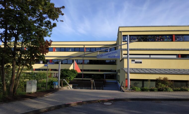 Schulgebäude mit Eingang und Flagge im Vordergrund, blauer Himmel im Hintergrund.