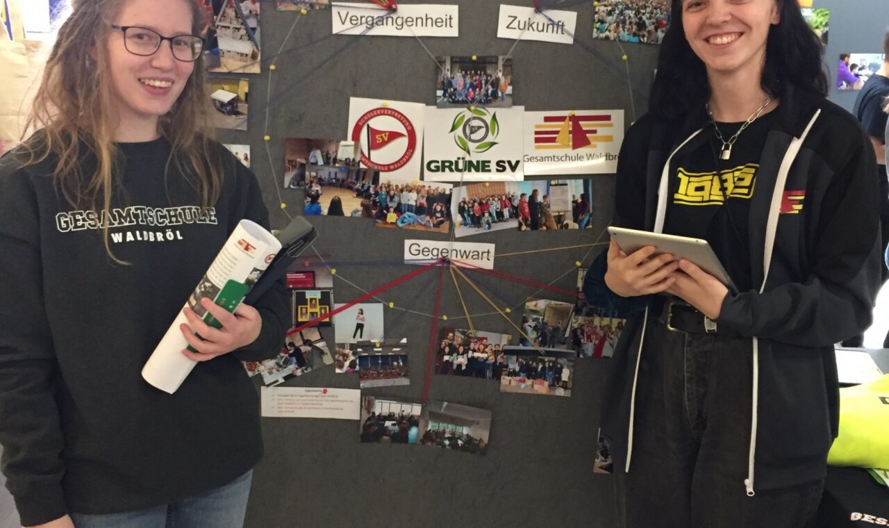 Zwei Personen stehen vor einem Plakat mit Fotos und Logos von Veranstaltungen.
