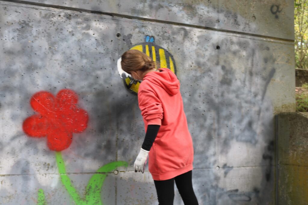 Person sprayt ein Graffiti mit einer Biene und einer roten Blume an einer Wand.